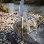Hand forged damascus viking bowie knife with a rosewood handle and rune cut brass bolster standing point down on river pebbles beside its brown leather sheath