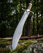 Hand forged Damascus Viking long seax with broken back blade and brass guard, low angle view upright in forest with its sheath