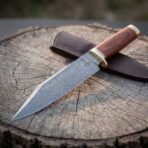 Hand forged Damascus Viking seax with rosewood handle, close view of the patterned broken back blade on a stump by its sheath