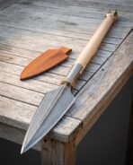 Hand forged damascus viking spear with a broad leaf blade, brass collar and riveted steel socket on an ash shaft, lying on a weathered table beside its leather blade cover
