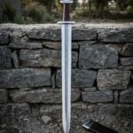 Hand forged damascus viking sword with a serpentine pattern welded core and Valknut inlaid lobed pommel standing point down against a dry stone wall at dusk