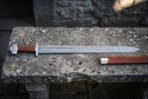 Hand forged damascus viking sword with a wide fullered blade, five lobed steel pommel and brown leather wrapped grip lying on a lichen covered stone bench beside its leather scabbard