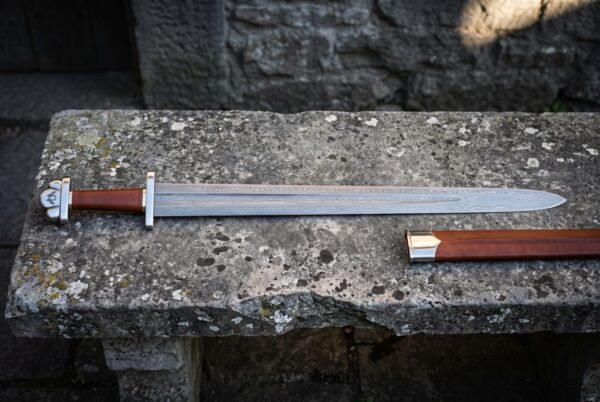 Hand forged damascus viking sword with a wide fullered blade, five lobed steel pommel and brown leather wrapped grip lying on a lichen covered stone bench beside its leather scabbard