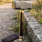 Hand forged Damascus viking war hammer standing upright on gravel, etched layered steel head with rosewood haft, brass bolsters and pommel, leather cover beside it