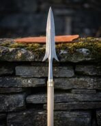 Hand forged Damascus winged spear with swept lugs, brass ferrule and pale ash haft standing against a mossy dry stone wall beside a tan leather head sheath