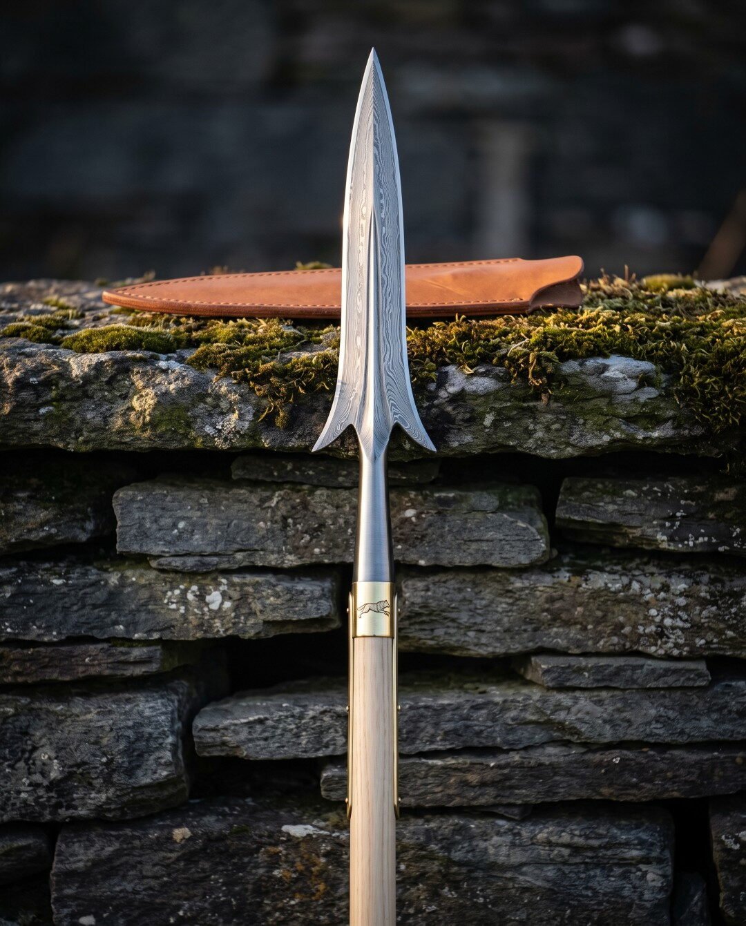 Hand forged Damascus winged spear with swept lugs, brass ferrule and pale ash haft standing against a mossy dry stone wa Hand forged Damascus winged spear with swept lugs, brass ferrule and pale ash haft standing against a mossy dry stone wall beside a tan leather head sheath