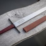 Hand forged display sword on a pale wooden bench, blunt rounded point and serpent etched blade, polished wheel pommel and burgundy leather grip with a brown scabbard