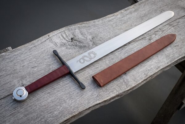 Hand forged display sword on a pale wooden bench, blunt rounded point and serpent etched blade, polished wheel pommel and burgundy leather grip with a brown scabbard