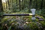 Hand forged double bit axe in 1095 steel with an etched knotwork head, brass collar and long dark hardwood haft, resting on a mossy boulder in woodland