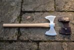 Hand forged double bit axe with etched Celtic knotwork on both flared bits and a pale ash haft, laid on mossy flagstones beside a brown leather head cover.
