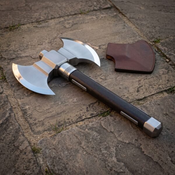 Hand forged double headed battle axe with polished twin crescent bits, engraved knotwork collar and dark hardwood haft on flagstones beside a leather edge cover