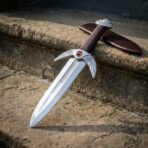 Hand forged fantasy dagger with winged guard and red cabochon leaning on a stone step, close view of the blade and sheath