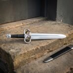 Hand forged fantasy sword with a twisted loop guard, black leather grip and polished steel wheel pommel, lying on a stone step beside its black scabbard with a steel chape.