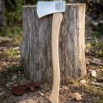 Hand forged felling axe with a polished 1095 steel head, knotwork and rune engraving and a natural ash handle leaning against a cut stump in woodland