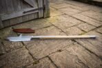 Hand forged glaive polearm with a broad hooked single edged head and knotwork engraved socket on a long ash haft, lying on flagstones beside a brown leather sheath.