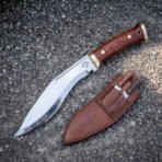 Hand forged Gurkha kukri knife with rosewood scales and etched wolf head, on gravel beside the sheath holding two companion knives