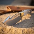 Hand forged gut hook hunting knife with stag antler handle, low close view of the hook and etched blade on sandstone