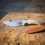 Hand forged gut hook knife with a mirror polished 1095 blade, etched runes, brass bolster and stag antler handle, on desert sandstone beside a tan leather sheath.