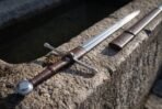 Hand forged high carbon hand and a half sword, close view of the round pommel, leather grip and cross guard on stone