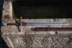 Hand forged high carbon hand and a half sword shown full length on a stone water trough above its brown leather scabbard