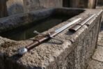 Hand forged high carbon hand and a half sword with brown leather grip lying on a stone trough beside its leather scabbard