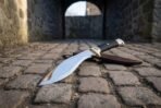Hand forged high carbon kukri with black grip and brass bolster, angled on cobblestones in a stone archway with its leather sheath