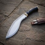 Hand forged high carbon steel kukri knife with black horn handle and brass bolster, rune etched blade on flagstones beside its sheath