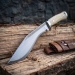 Hand forged high carbon steel kukri knife with bone handle scales and steel bolster, on a split log beside its leather sheath