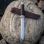 Hand forged high carbon tanto knife with stacked leather handle, lying point down on its leather sheath across a granite boulder