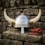 Hand forged horned Viking helmet with a riveted steel dome, brass rivets and trim, engraved knotwork browband, nasal bar and pale horns, on a dry stone wall.