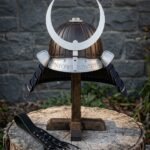 Hand forged kabuto helmet with a ribbed bronze bowl, polished steel crescent crest and rune cut brow band on a wooden stand before a slate wall.