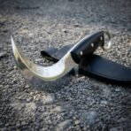 Hand forged karambit with black micarta scales and polished hooked blade resting across its black leather sheath on gravel