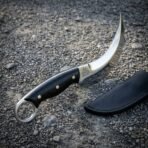 Hand forged karambit knife with curved blade, black micarta scales and ring pommel on gravel beside a black leather sheath