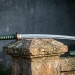 Hand forged katana sword with polished curved blade, green cord wrapped handle and dark iron tsuba resting on a weathered stone pillar beside its green saya