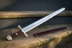 Hand forged knightly sword with a polished fullered blade, engraved knotwork panel, brass crossguard and pommel and oxblood leather grip, resting on a stone step beside its burgundy scabbard.