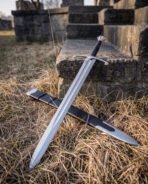 Hand forged knotwork longsword leaning across a low stone step in dry grass with its black leather scabbard and polished steel chape crossing below