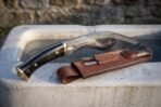 Hand forged kukri with horn handle and brass pommel lying on stone, tan leather sheath holding two small knives in front