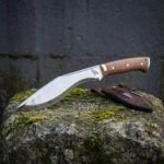 Hand forged kukri knife in mirror polished high carbon steel with walnut scales, brass bolster and a raven etch, on mossy stone beside its leather sheath and companion knives