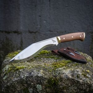 Hand forged kukri knife in mirror polished high carbon steel with walnut scales, brass bolster and a raven etch, on mossy stone beside its leather sheath and companion knives