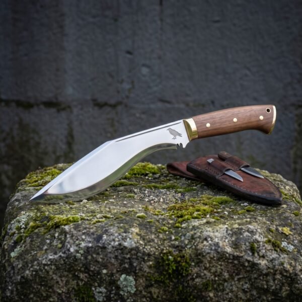 Hand forged kukri knife in mirror polished high carbon steel with walnut scales, brass bolster and a raven etch, on mossy stone beside its leather sheath and companion knives