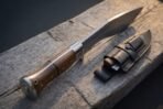 Hand forged kukri knife with rosewood scales and steel bolster resting on a timber beam beside a leather sheath with companion blades