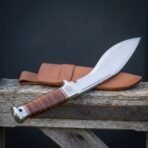 Hand forged kukri knife with stacked leather handle, handle in the foreground and blade rising over a wooden rail beside its sheath