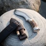 Hand forged kukri knife with stag horn scales and steel bolster on a pale rock beside a leather belt sheath and small blades