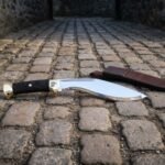 Hand forged kukri sword lying on cobblestones, mirror polished deep bellied blade, black cord wrapped grip and a raven engraved brass bolster with leather sheath