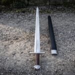 Hand forged leather grip viking sword laid on grey gravel with a lobed pommel, an etched knotwork panel above the guard and a black leather scabbard alongside