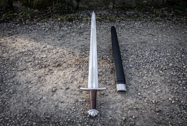 Hand forged leather grip viking sword laid on grey gravel with a lobed pommel, an etched knotwork panel above the guard and a black leather scabbard alongside