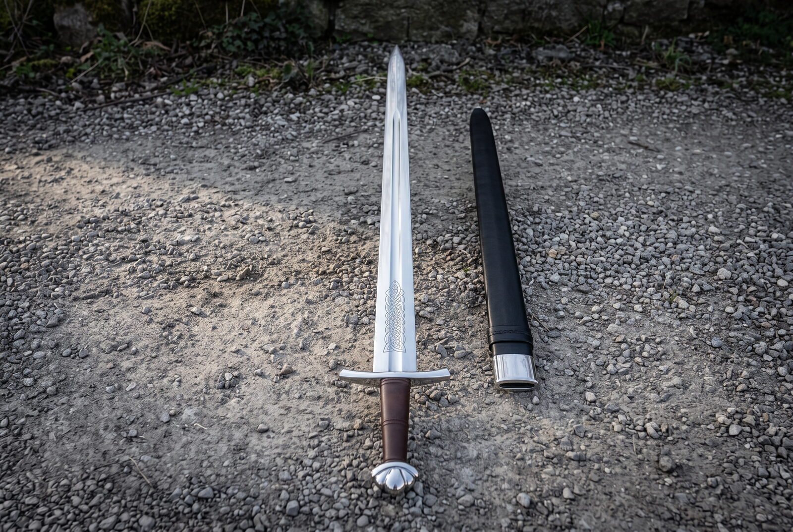 Hand forged leather grip viking sword laid on grey gravel with a lobed pommel, an etched knotwork panel above the guard Hand forged leather grip viking sword laid on grey gravel with a lobed pommel, an etched knotwork panel above the guard and a black leather scabbard alongside
