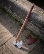 Hand forged long handled Viking axe with leather grip wrap, leaning against a castle wall walk beside its fitted leather blade cover