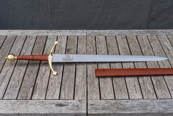 Hand forged longship sword laid on weathered decking, brass ball pommel and trefoil crossguard, tan leather grip and a Norse ship etched on the polished blade