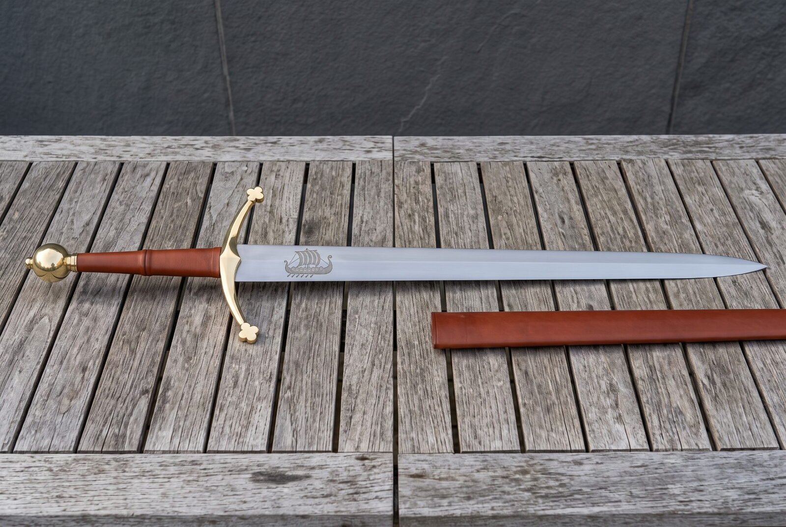 Hand forged longship sword laid on weathered decking, brass ball pommel and trefoil crossguard, tan leather grip and a N Hand forged longship sword laid on weathered decking, brass ball pommel and trefoil crossguard, tan leather grip and a Norse ship etched on the polished blade
